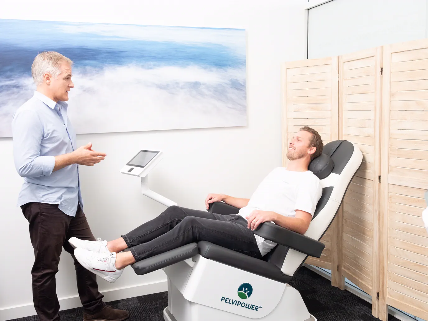 Nathan talking with a man seated, fully clothed, on the PelviPower chair in the CoreGood clinic